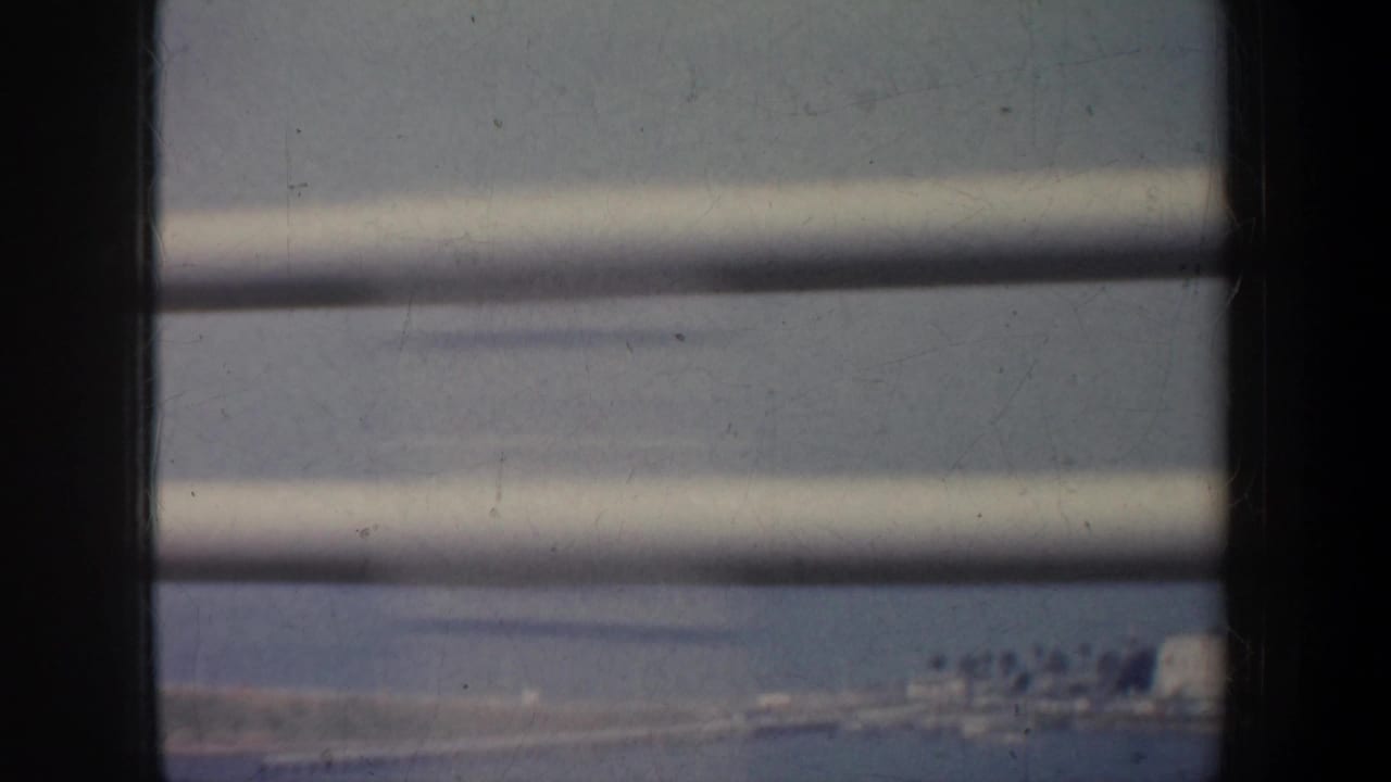 1957 San Francisco home-movie frame looking through horizontal blinds toward a soft, distant landscape.