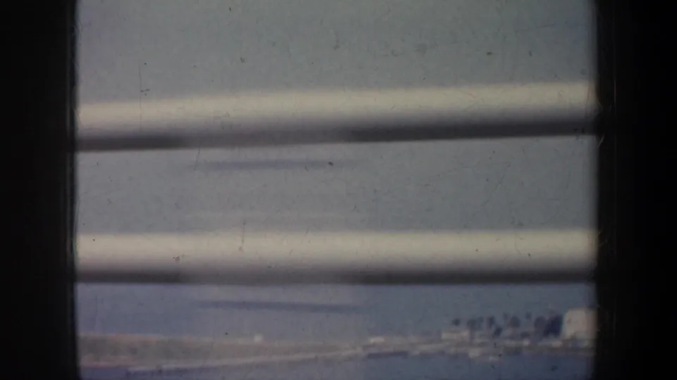 1957 San Francisco home-movie frame looking through horizontal blinds toward a soft, distant landscape.