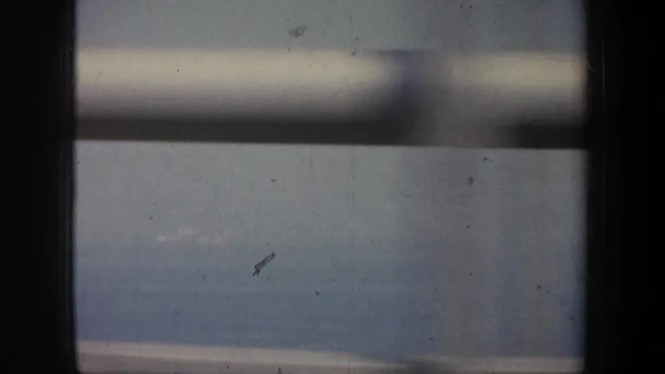 1957 San Francisco home-movie frame looking through bridge structure toward blue-gray water and a faint shoreline.