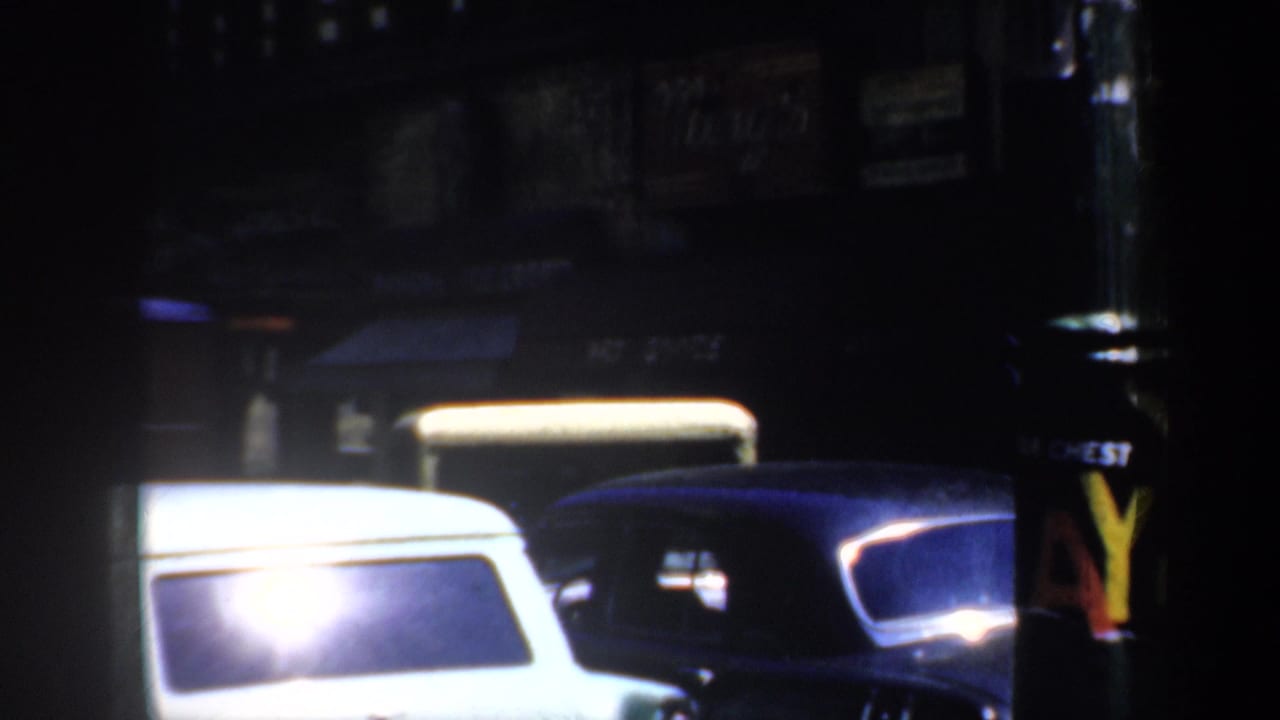 1957 San Francisco street at night with parked mid-century cars, storefront awnings, and bright reflections in the film frame.