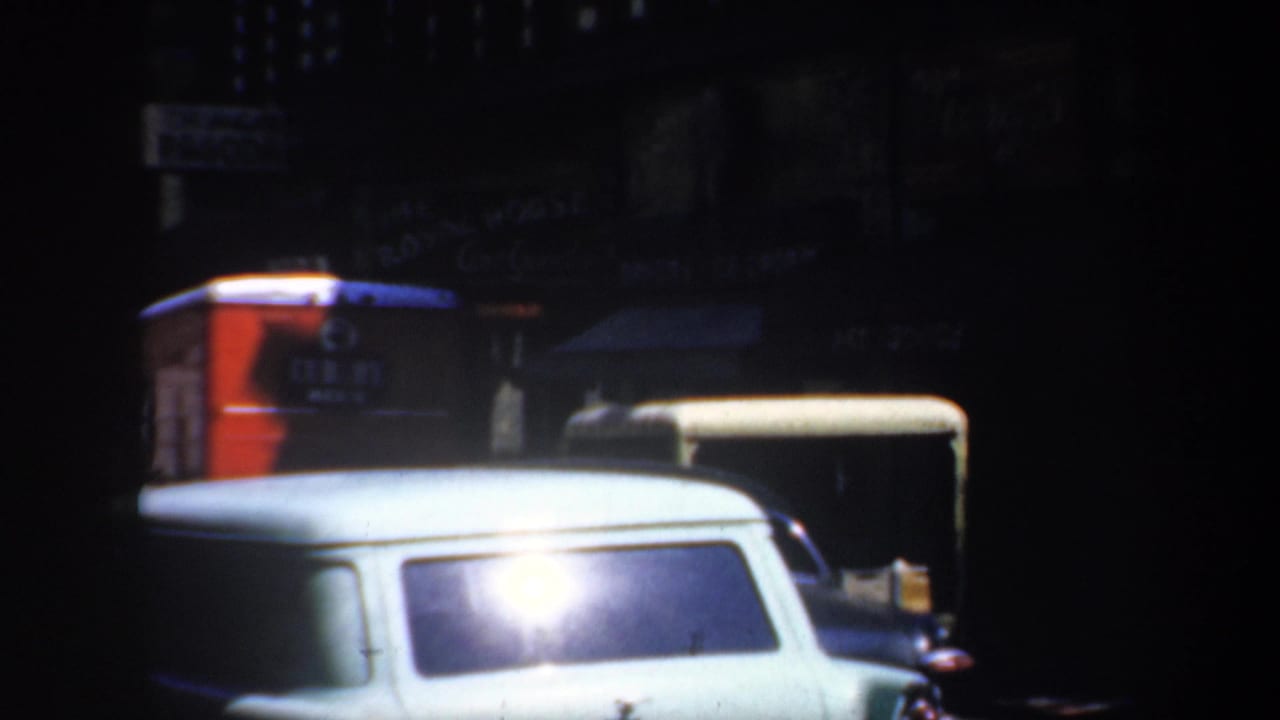 Nighttime street frame with a pale station wagon, red sign, awnings, and dark parked cars.