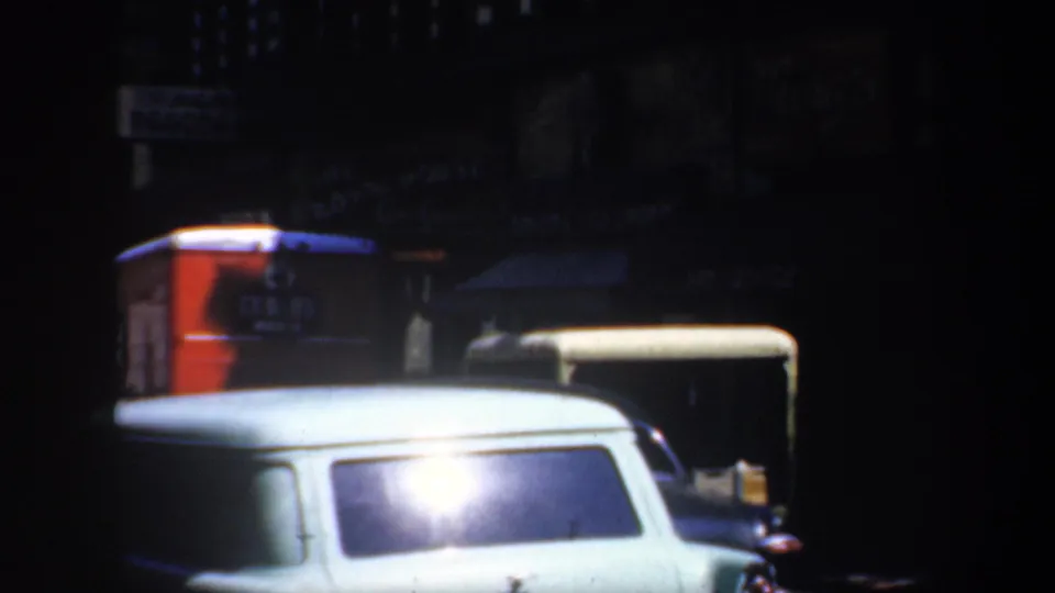 Night view of a San Francisco commercial street with a white station wagon, dark vehicles, awnings, and a red storefront sign.