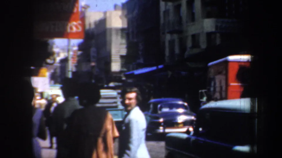 Daytime San Francisco street scene with pedestrians walking near parked 1950s cars and storefront signage on a commercial block.