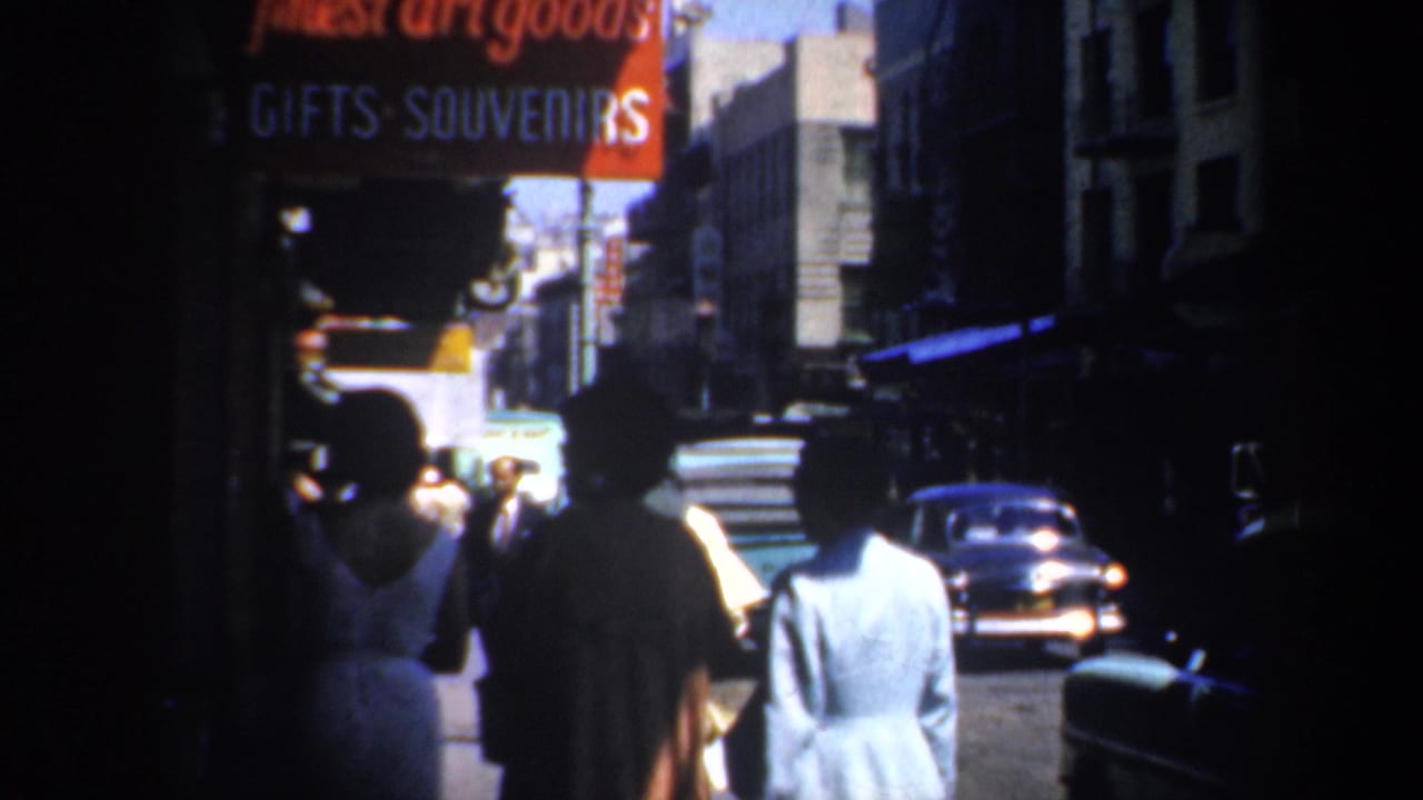 1957 San Francisco street view with pedestrians from behind, souvenir signage, deep sidewalk shadows, and a mid-century car ahead.