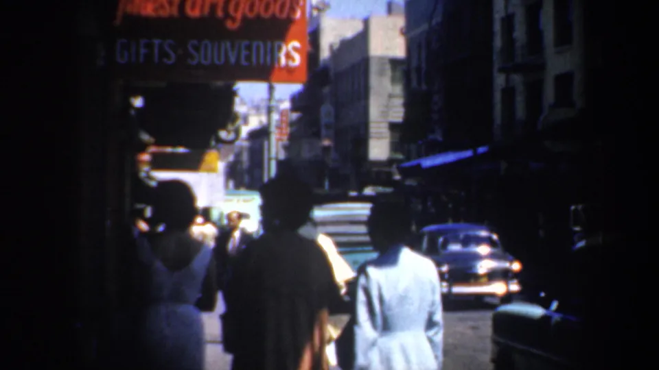 1957 San Francisco street view with pedestrians from behind, souvenir signage, deep sidewalk shadows, and a mid-century car ahead.
