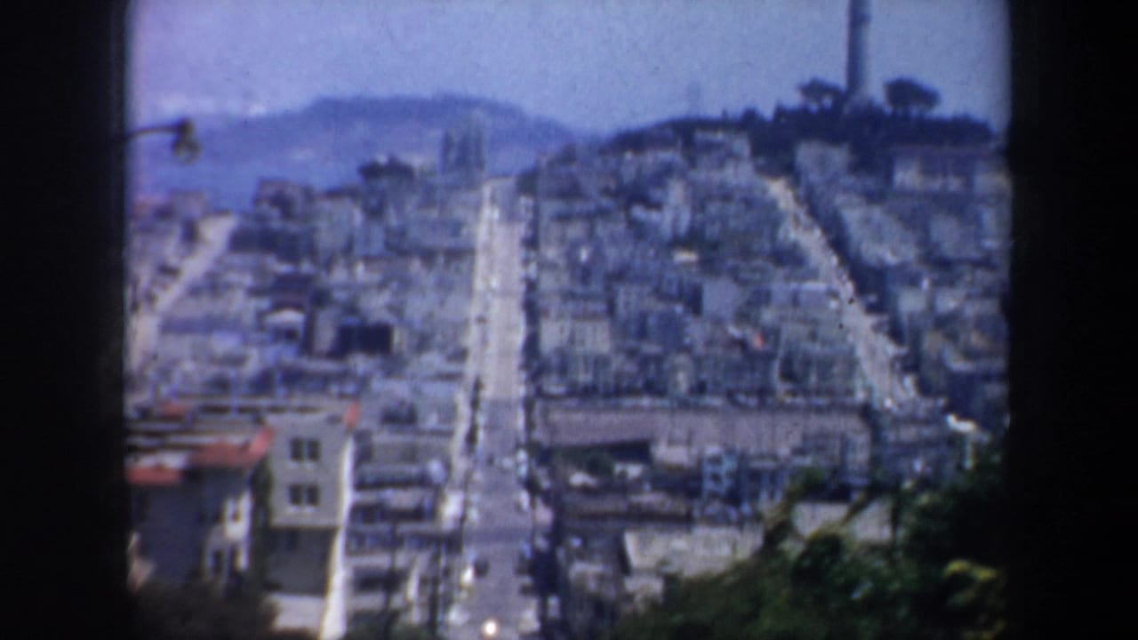 Telephoto-like city view down a steep street toward bay water and a distant hillside.