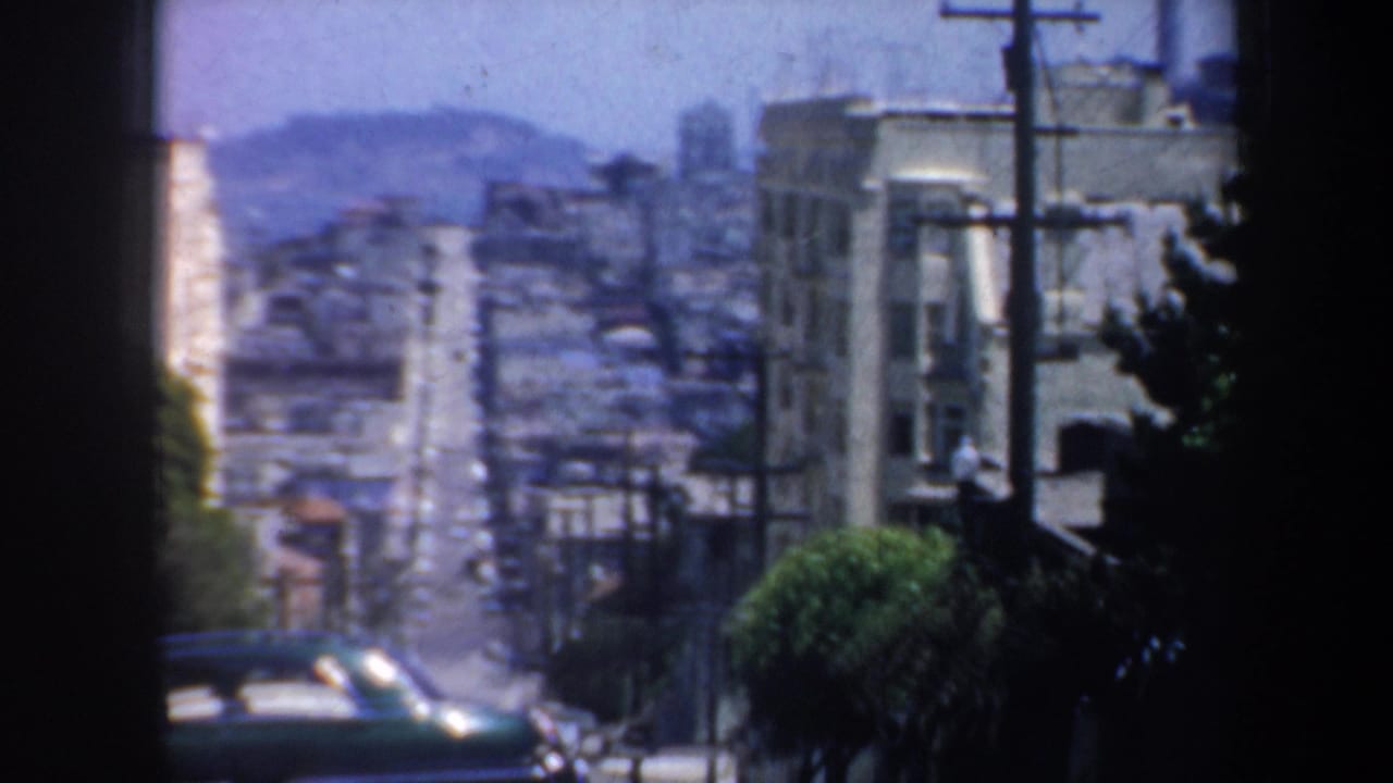 1957 San Francisco street view from a moving vehicle, showing a steep grade, mid-century cars, utility poles, and dense buildings.