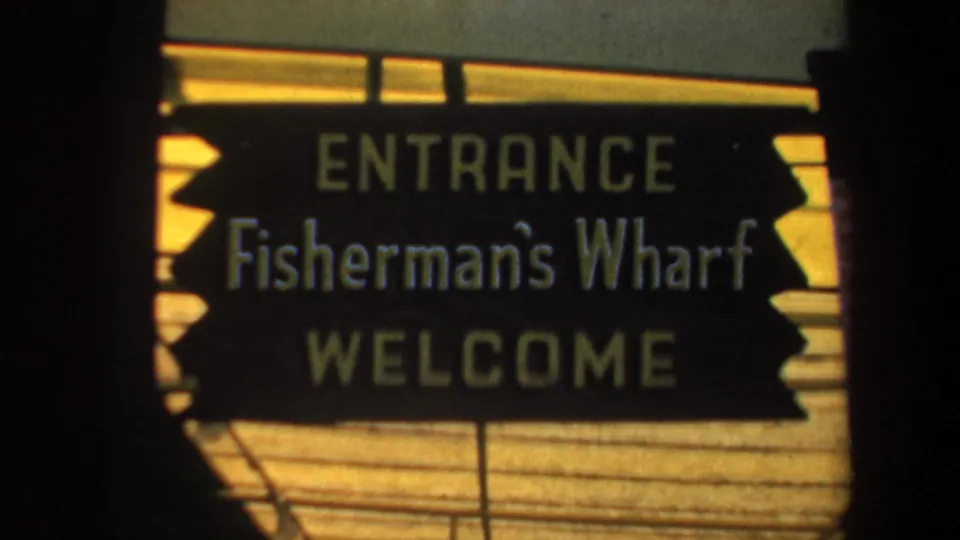 Tighter 1957 home-movie view of the Fisherman's Wharf entrance sign, framed with dark film edges and warm sunlight.