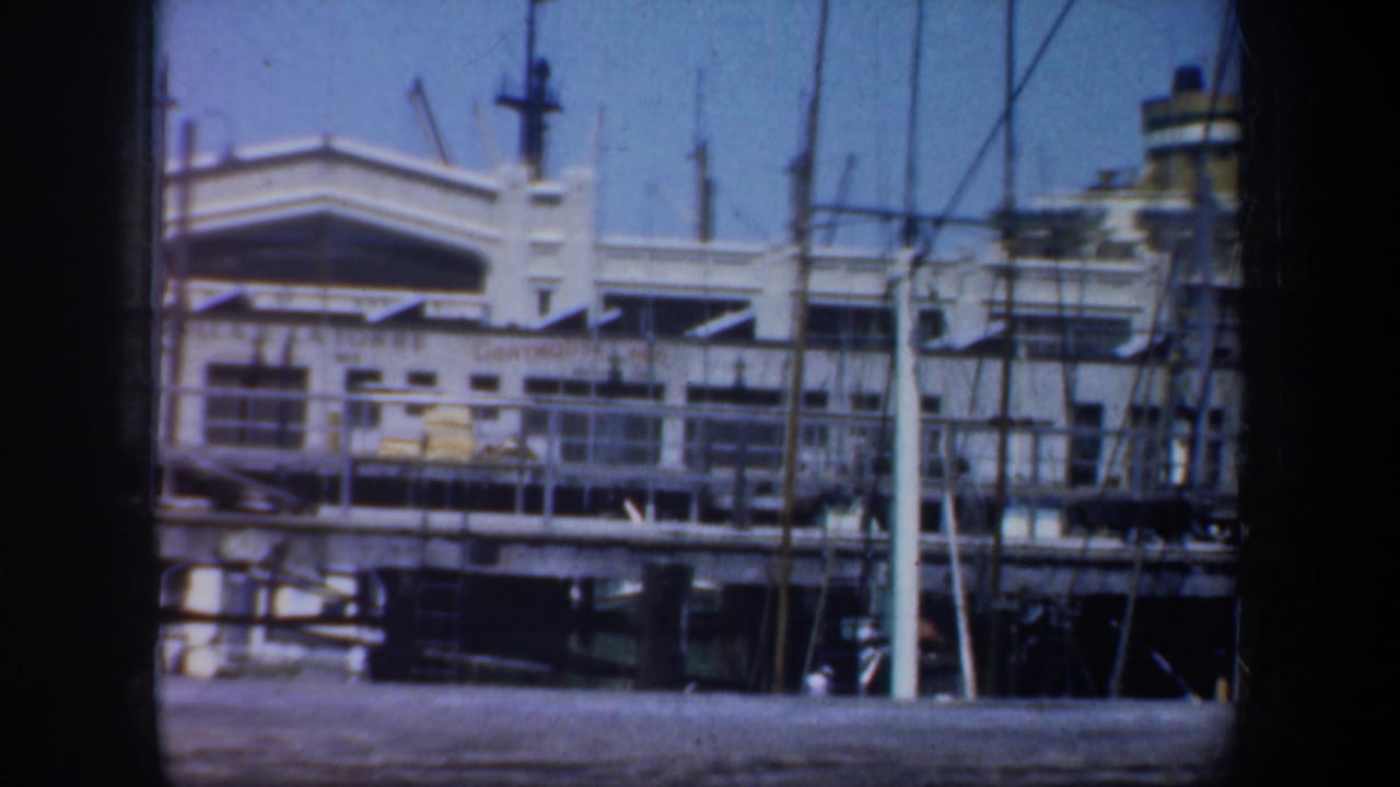 White waterfront building seen across dock railings with sailboat masts in front.