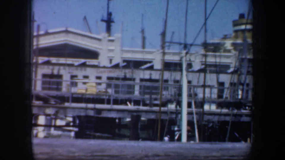 1957 San Francisco waterfront view with a white pier-side building, dock railing, masts, and harbor water in the foreground.