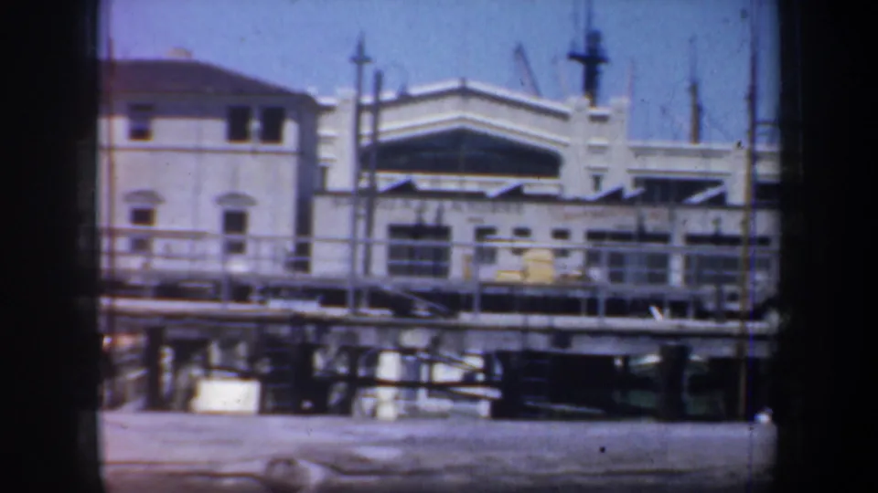1957 San Francisco home-movie frame showing dock railings, a white waterfront structure, masts, windows, and harbor water.