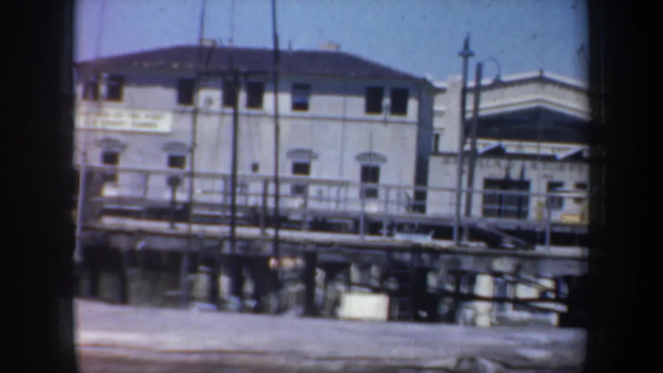 1957 San Francisco waterfront frame with white pier buildings, dock railing, vertical masts, harbor water, and sunlit sky.