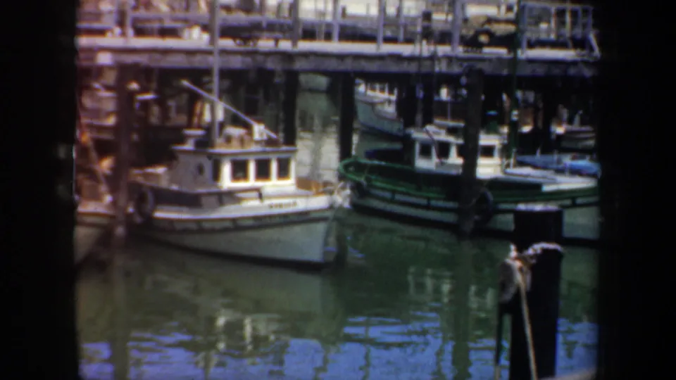 1957 San Francisco dockyard frame showing moored small boats, pilings, pier railings, and calm green harbor water.