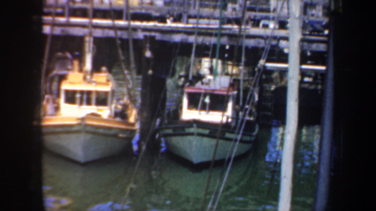 1957 San Francisco harbor frame showing two small fishing boats tied up beneath a wooden pier, with masts, rigging, and calm green water visible.