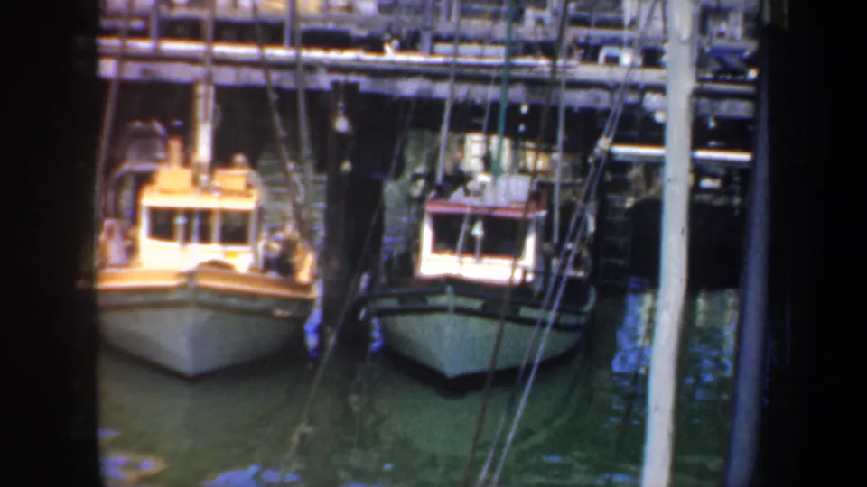 1957 San Francisco harbor frame showing two small fishing boats tied up beneath a wooden pier, with masts, rigging, and calm green water visible.