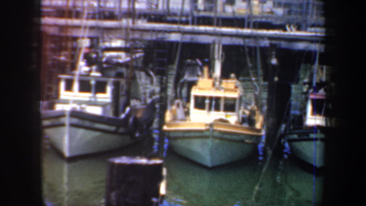 1957 San Francisco harbor view of small fishing boats moored alongside pilings and wooden dock structures, with rigging and industrial waterfront details behind them.