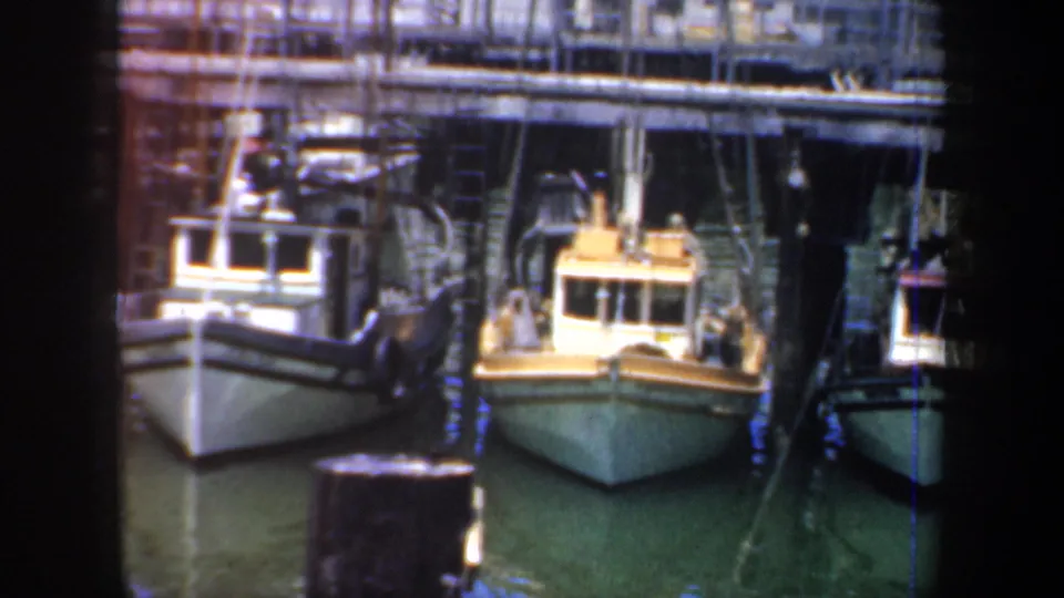 1957 San Francisco harbor view of small fishing boats moored alongside pilings and wooden dock structures, with rigging and industrial waterfront details behind them.