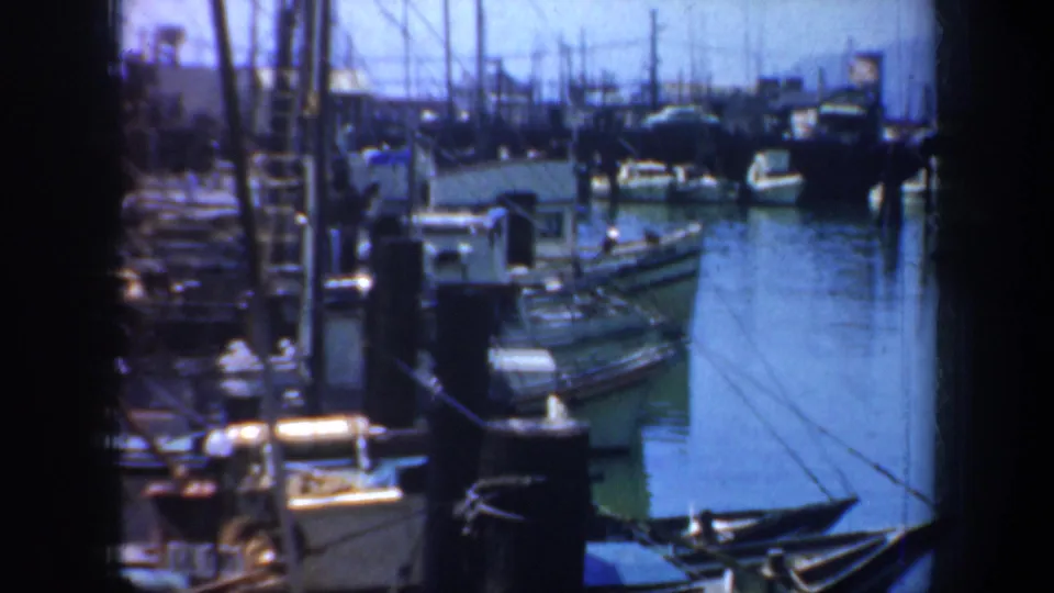 1957 San Francisco harbor scene with small boats at rest beside pilings, visible rigging, dock equipment, and calm water in a soft 8mm home-movie frame.