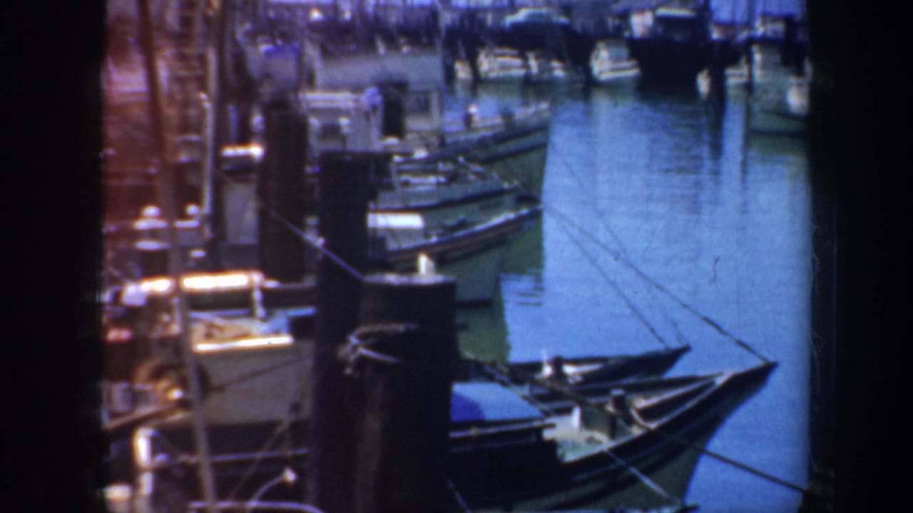 1957 San Francisco harbor footage showing small boats tied near a wharf, blue water, ropes, and dock structures in a soft 8mm frame.
