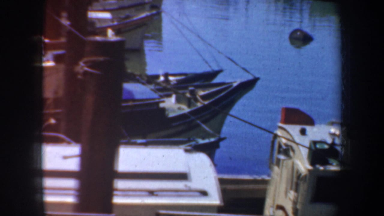 1957 San Francisco waterfront view with small boat bows, mooring lines, dock structures, and calm blue water captured on 8mm film.