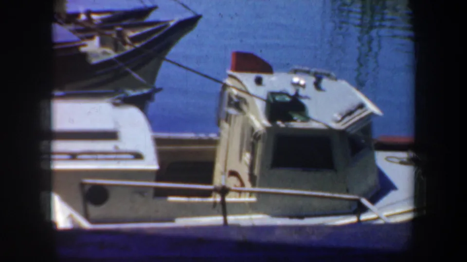 1957 San Francisco harbor frame showing a small cabin boat at the dock, nearby boat bow, railings, ropes, and bright blue water.