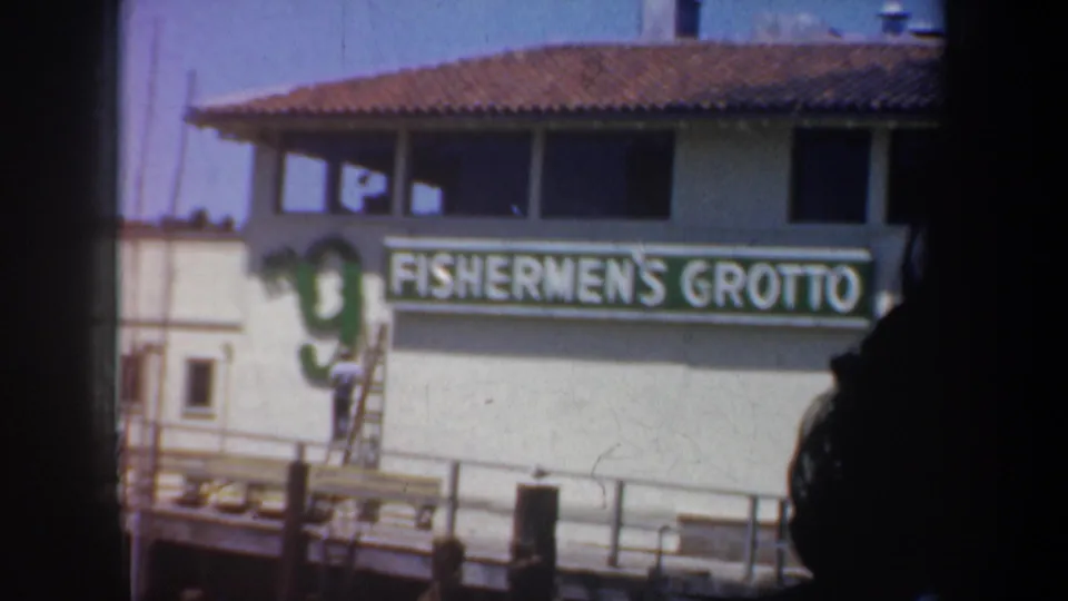 1957 San Francisco harbor frame showing the Fishermen's Grotto sign, dock railings, a ladder, and a worker near the waterfront building.