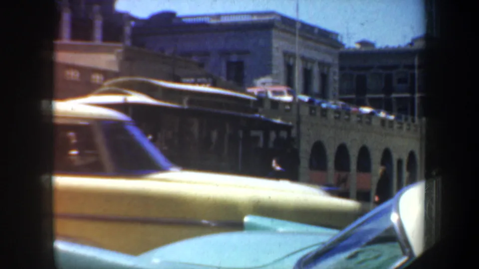 1957 San Francisco street frame with a yellow mid-century car passing a trolley and a teal vehicle hood in the foreground.
