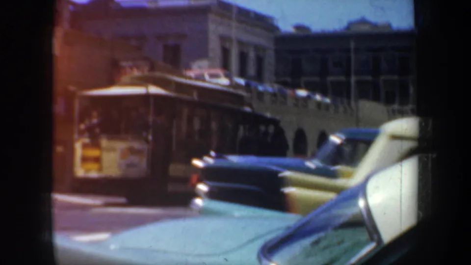 1957 San Francisco 8mm frame with a trolley, yellow and dark cars, and a teal vehicle foreground against older buildings.