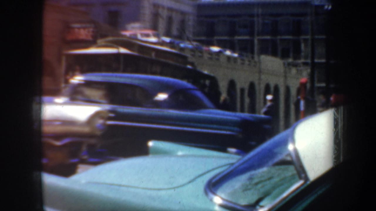 Downtown street with turquoise car hood, passing sedan, and yellow transit vehicle.