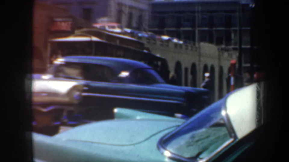 1957 San Francisco street view with a turquoise car in the foreground, a dark sedan passing, and a yellow transit vehicle behind it.