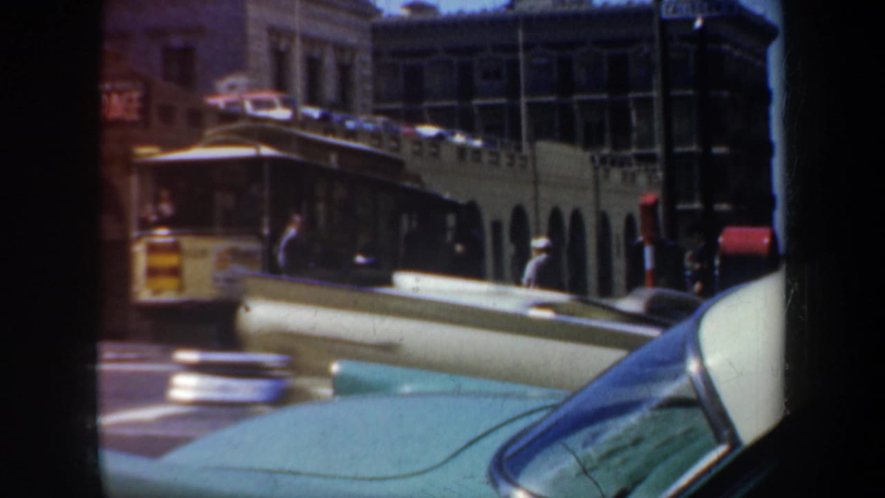 San Francisco street traffic in 1957, with a yellow transit vehicle moving through the intersection behind a turquoise car hood.