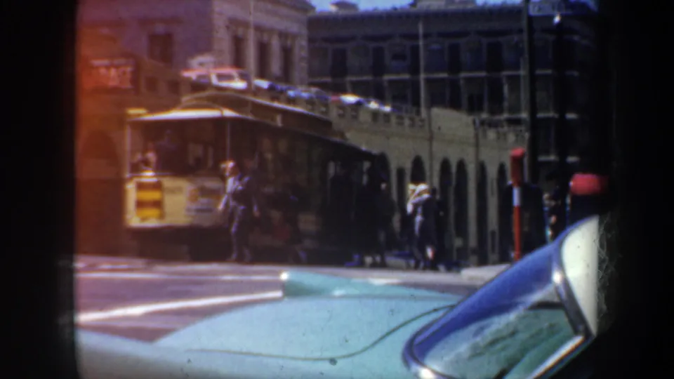 1957 San Francisco downtown scene with a transit vehicle near the curb, pedestrians on the sidewalk, and a turquoise car in the foreground.