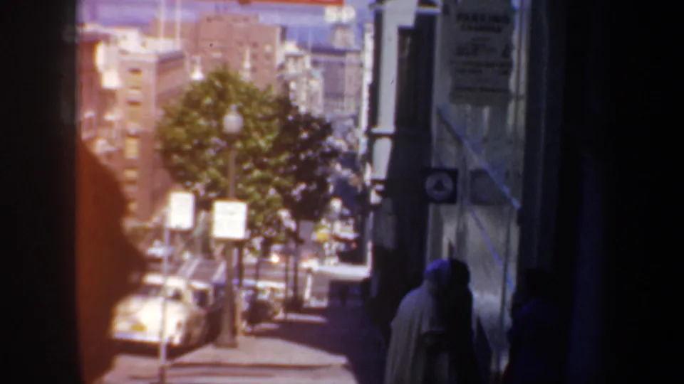 San Francisco sidewalk view from 1957 with parked cars, street trees, posted signs, and pedestrians walking beside a building edge.
