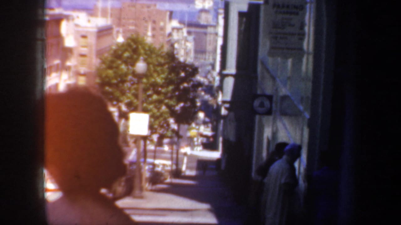 Sunlit city sidewalk with pedestrians, buildings, trees, and street activity.