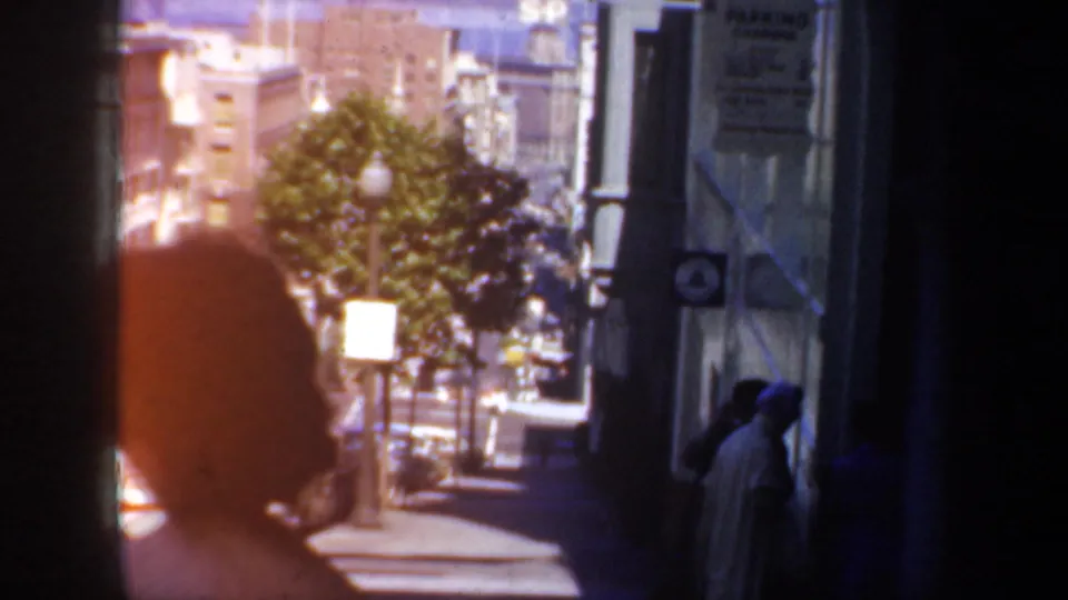 1957 San Francisco home-movie view of a sunlit sidewalk with pedestrians, storefronts or building fronts, trees, and street traffic beyond.