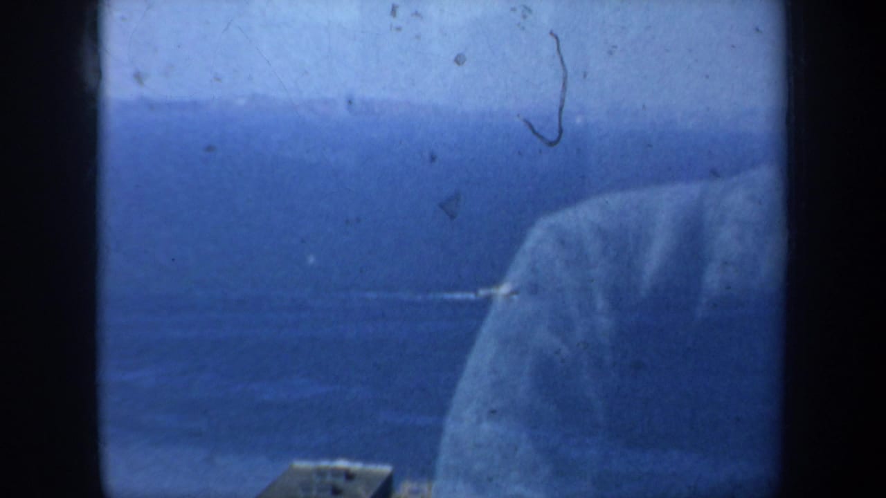 Elevated 1957 San Francisco bay view in blue-shifted 8mm color, with water, a shoreline edge, and a large pale form near the right side.