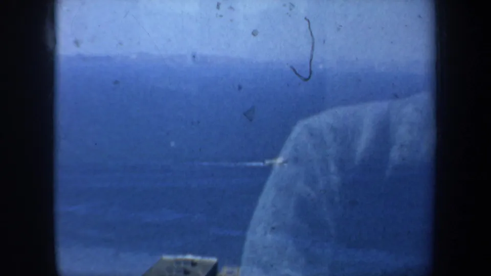 Elevated 1957 San Francisco bay view in blue-shifted 8mm color, with water, a shoreline edge, and a large pale form near the right side.
