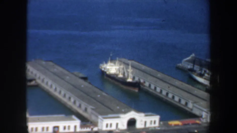 Elevated 1957 San Francisco waterfront view with white pier buildings, docked boats, long piers, blue bay water, and partial street edge.