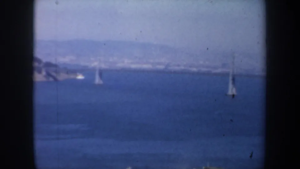 1957 San Francisco Bay view with distant Golden Gate Bridge towers and small sailboats in the foreground water.