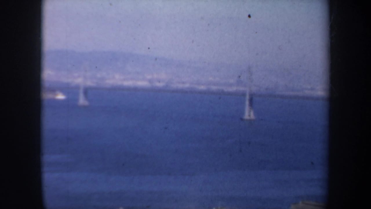 1957 San Francisco bay view with a distant bridge and small sailboats framed by dark vertical window edges.