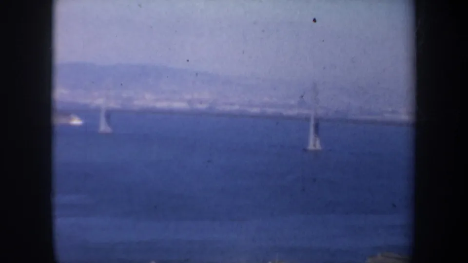 1957 San Francisco bay view with a distant bridge and small sailboats framed by dark vertical window edges.