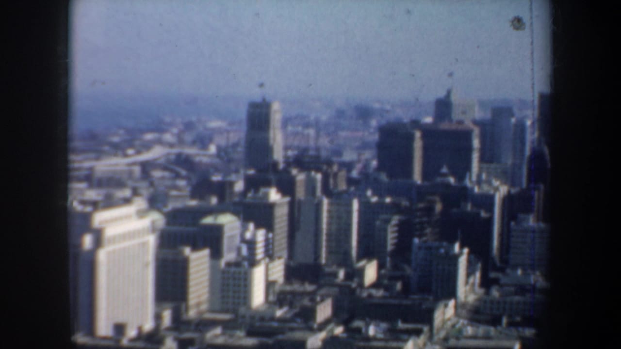 High-angle city view with towers, rooftops, and haze framed by dark window edges.