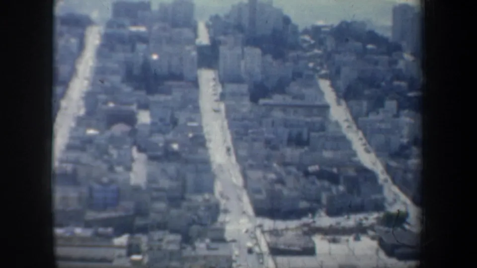 1957 San Francisco 8mm frame showing an elevated view of city streets, low-rise buildings, and distant hills through soft haze.