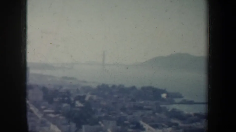 1957 San Francisco 8mm frame looking toward bay water, shoreline, faint bridge towers, and distant hills under a pale sky.