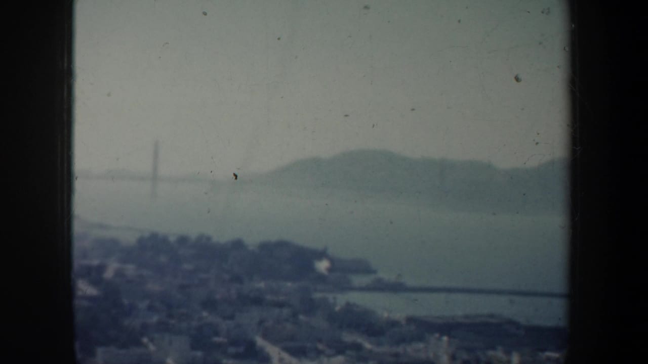 1957 San Francisco bay panorama through a window, with shoreline, distant hills, and a faint bridge or tower form in haze.