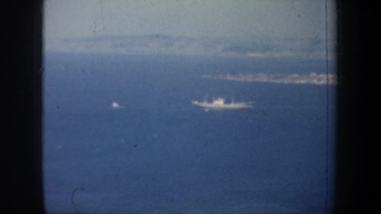 1957 San Francisco-area aerial view showing a distant ship on blue bay water beneath a hazy coastline and pale sky.