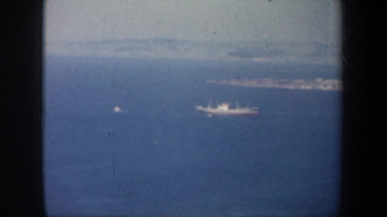 Aerial 8mm frame from the San Francisco bay area, with a large ship and smaller vessel set against haze and distant land.