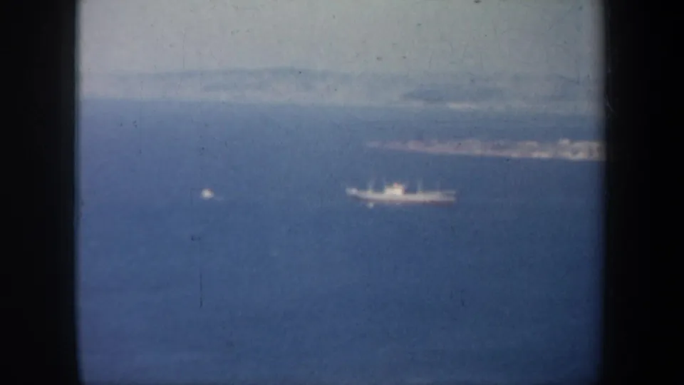 Aerial 8mm frame from the San Francisco bay area, with a large ship and smaller vessel set against haze and distant land.
