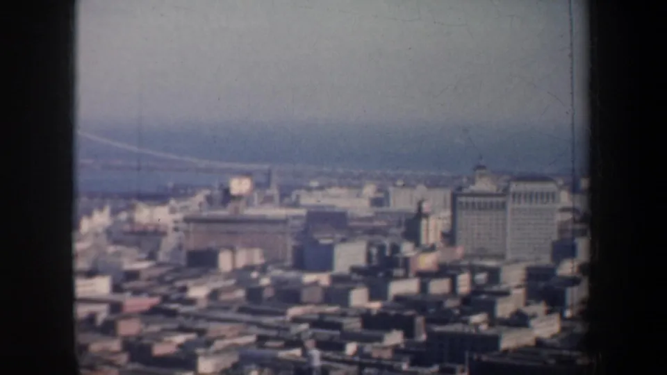 1957 aerial view over San Francisco rooftops and waterfront buildings, with bay water and the Golden Gate Bridge faintly visible beyond.