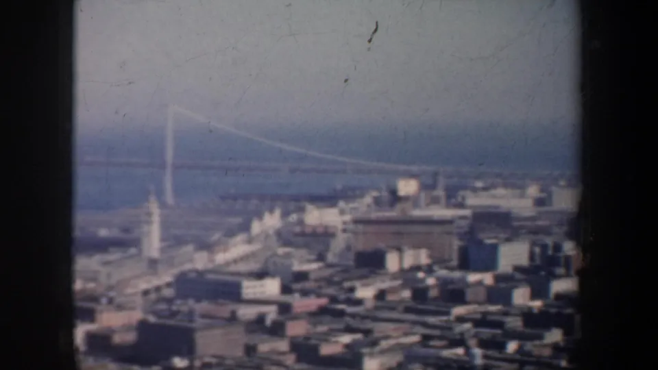 San Francisco aerial home-movie frame showing city rooftops, waterfront structures, bay haze, and the Golden Gate Bridge in the distance.